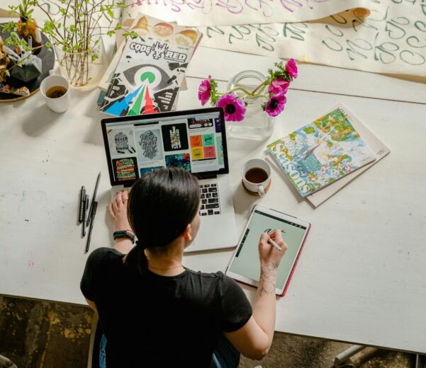 Person sitting at a white desk working on a laptop and tablet, surrounded by art supplies, colorful design prints, a vase of flowers, and coffee cups