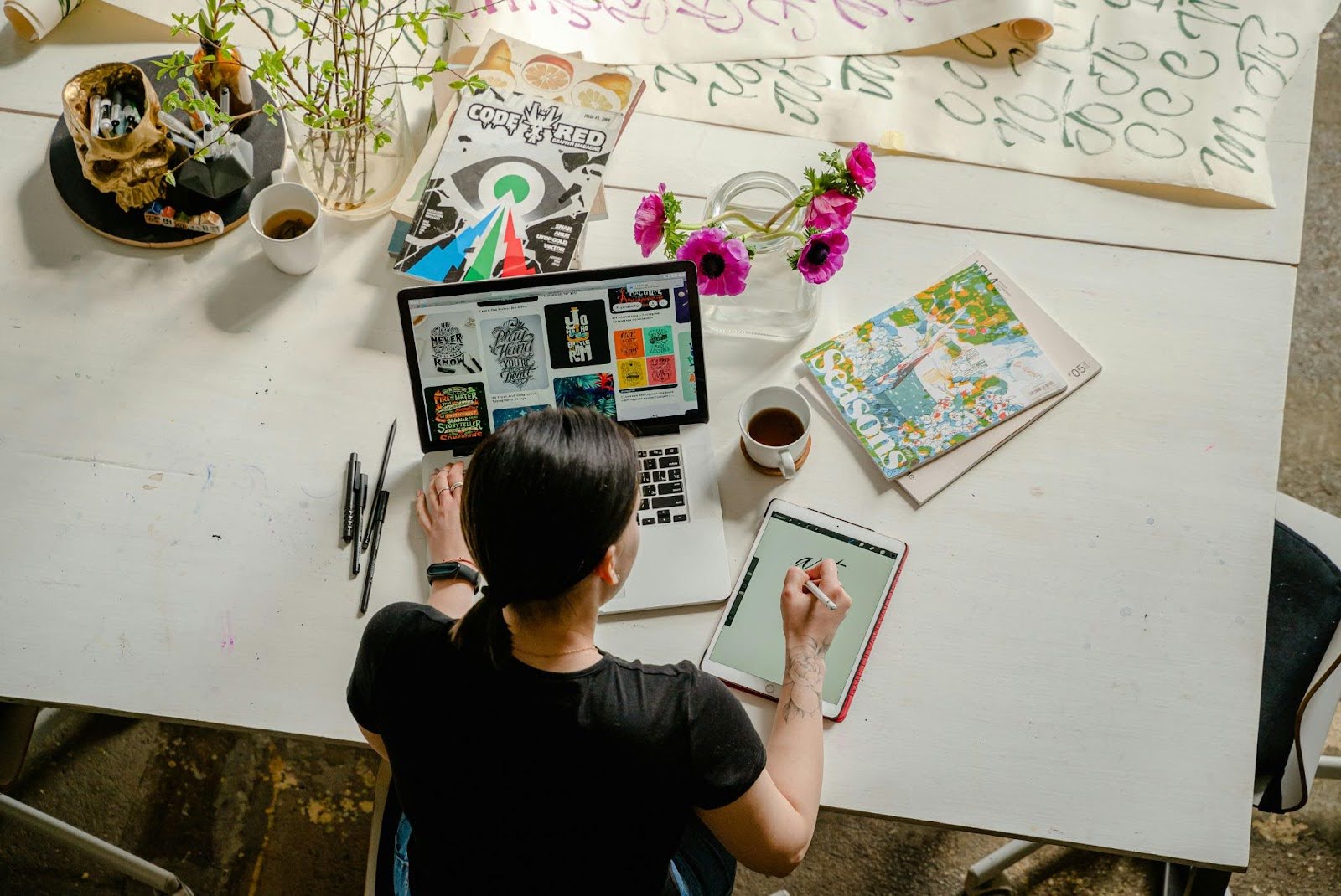 Person sitting at a white desk working on a laptop and tablet, surrounded by art supplies, colorful design prints, a vase of flowers, and coffee cups
